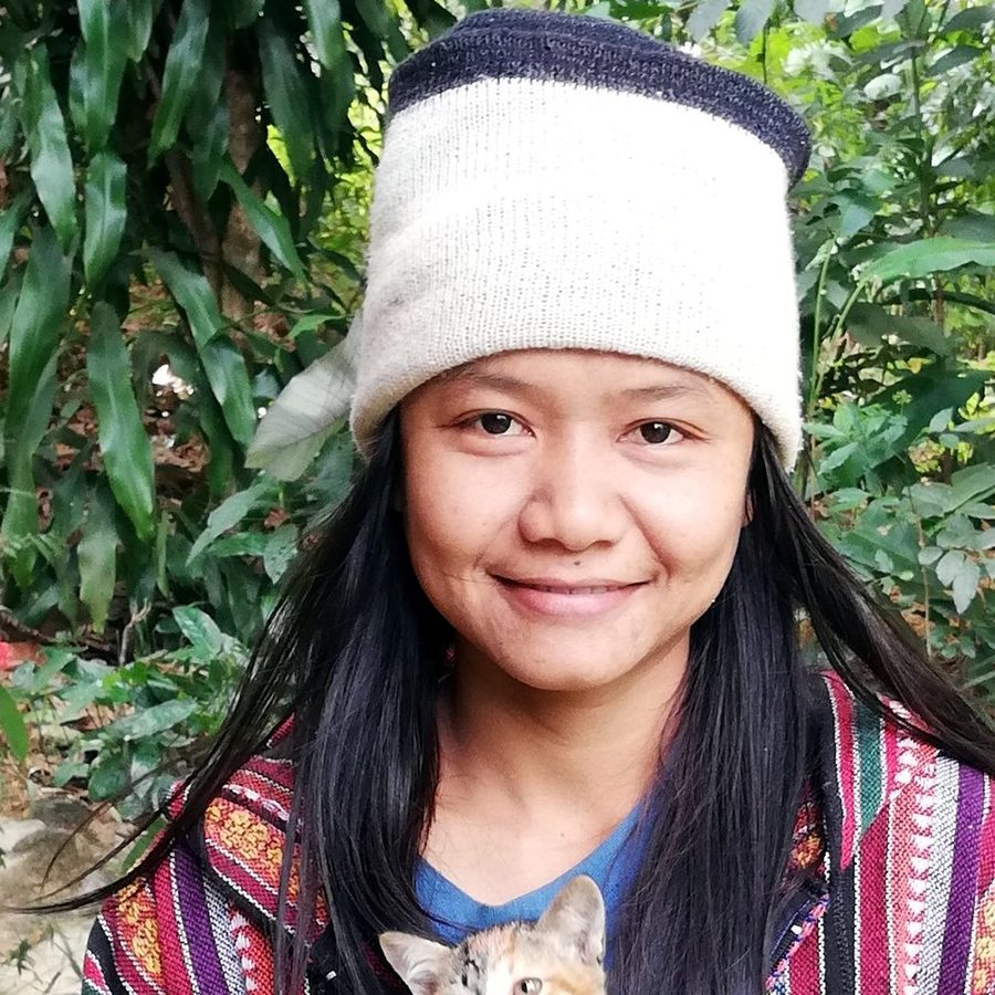 朴门实践者Anuthida "Karn" Moolnam 在绿树成荫的花园里抱着一只小猫，戴着针织帽和传统编织夹克，微笑着。