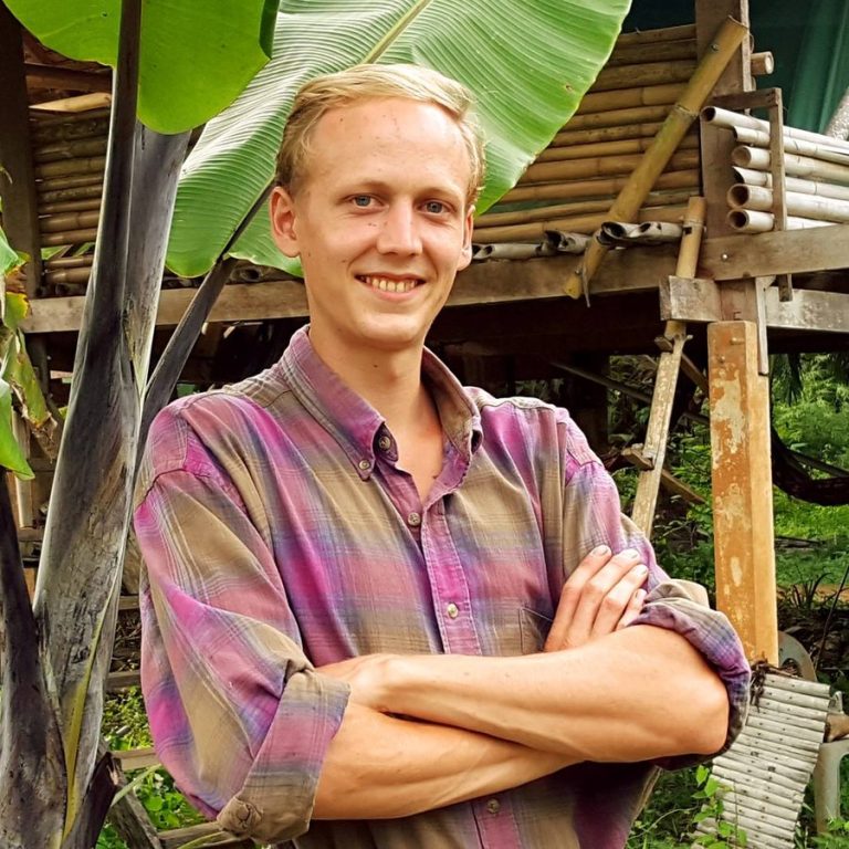 David B. Lauterwasser, permaculture designer, standing with arms crossed in front of a bamboo structure and banana leaf, Thailand.
