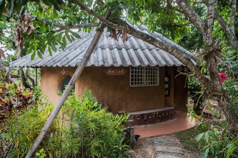 A small adobe earthen-brick guesthouse with a tiled hip roof and a Thai name plaque, nestled among trees and tropical plants beside a stone path, at Earth Home Thailand (Maejo Baandin), Mae Taeng, Chiang Mai.