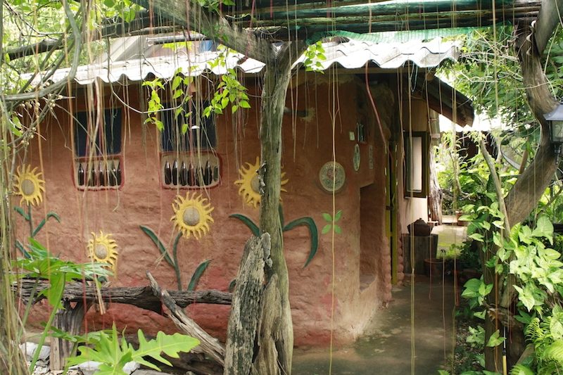 An adobe house with reddish earthen walls hand-painted with sunflowers and decorated with embedded bottles, glimpsed through hanging vines and greenery, at Earth Home Thailand (Maejo Baandin), Mae Taeng, Chiang Mai.