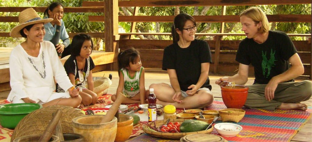 People sitting on a woven mat on a wooden deck around bowls, mortars and fresh ingredients during a Thai cooking session, including adults and children, at Earth Home Thailand (Maejo Baandin), Mae Taeng, Chiang Mai.