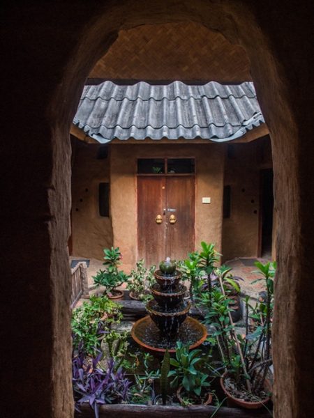 View through an arched earthen doorway into a planted inner courtyard with a tiered water fountain, potted plants, and a wooden door, at Earth Home Thailand (Maejo Baandin), Mae Taeng, Chiang Mai.