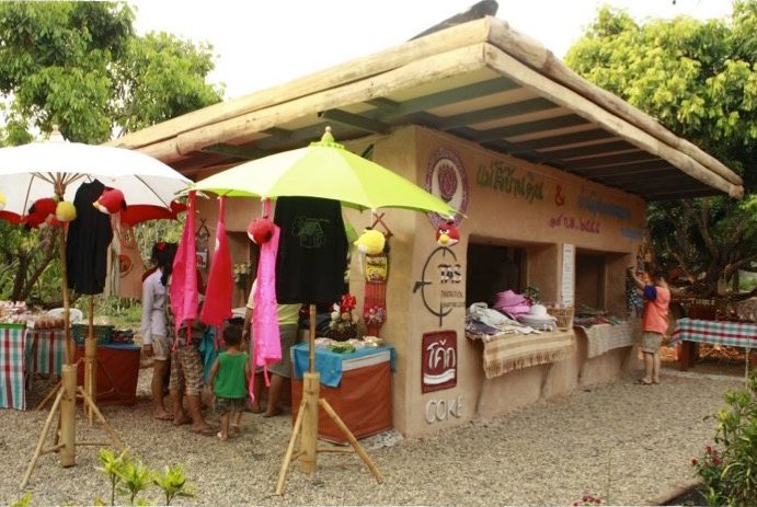 An open-sided earthen building used as a craft stall, with printed T-shirts and aprons hung under colourful parasols and people browsing, at Earth Home Thailand (Maejo Baandin), Mae Taeng, Chiang Mai.