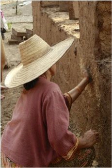A person in a straw hat smoothing wet earthen plaster onto a clay wall by hand, at Earth Home Thailand (Maejo Baandin), Mae Taeng, Chiang Mai.