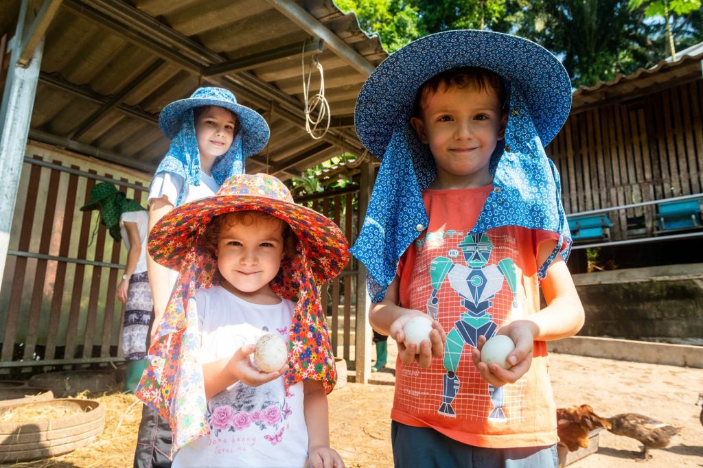 Three children in sun hats holding freshly collected eggs beside the chicken run on the farm at Eco-Logic Thailand, Phato, Chumphon.