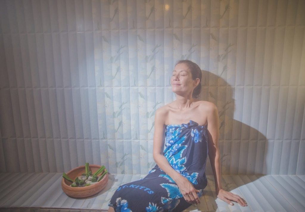 A woman relaxing with eyes closed beside a bowl of fresh herbs in the herbal steam sauna at Eco-Logic Thailand, Phato, Chumphon.