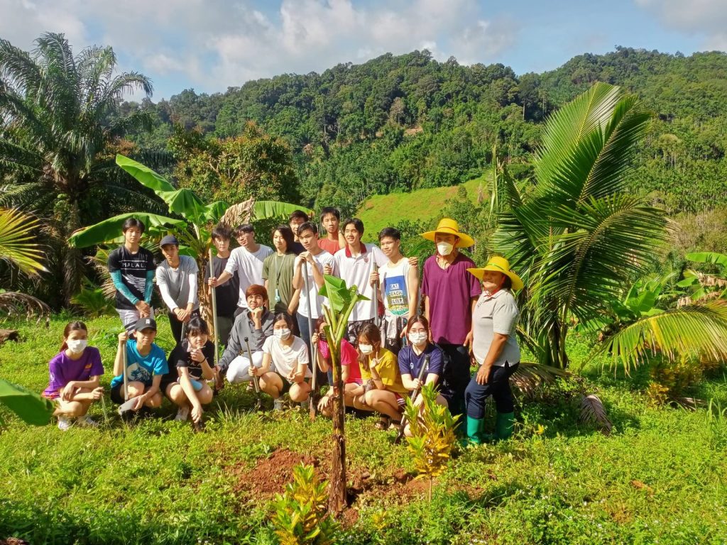 A group of young volunteers gathered around a newly planted tree on a green hillside with forest behind at Eco-Logic Thailand, Phato, Chumphon.