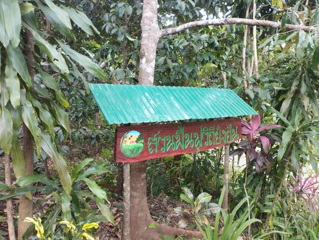 Hand-painted Thai-language entrance sign with a green corrugated-metal roof, mounted on wooden posts among tropical shrubs at the Feun Foo permaculture garden.