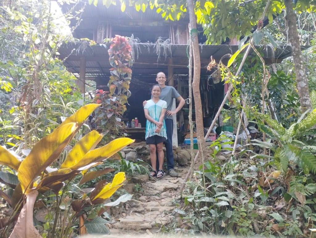 A man and a young girl standing arm in arm on a stone step in front of a rustic wooden cabin surrounded by dense tropical greenery at Feun Foo.