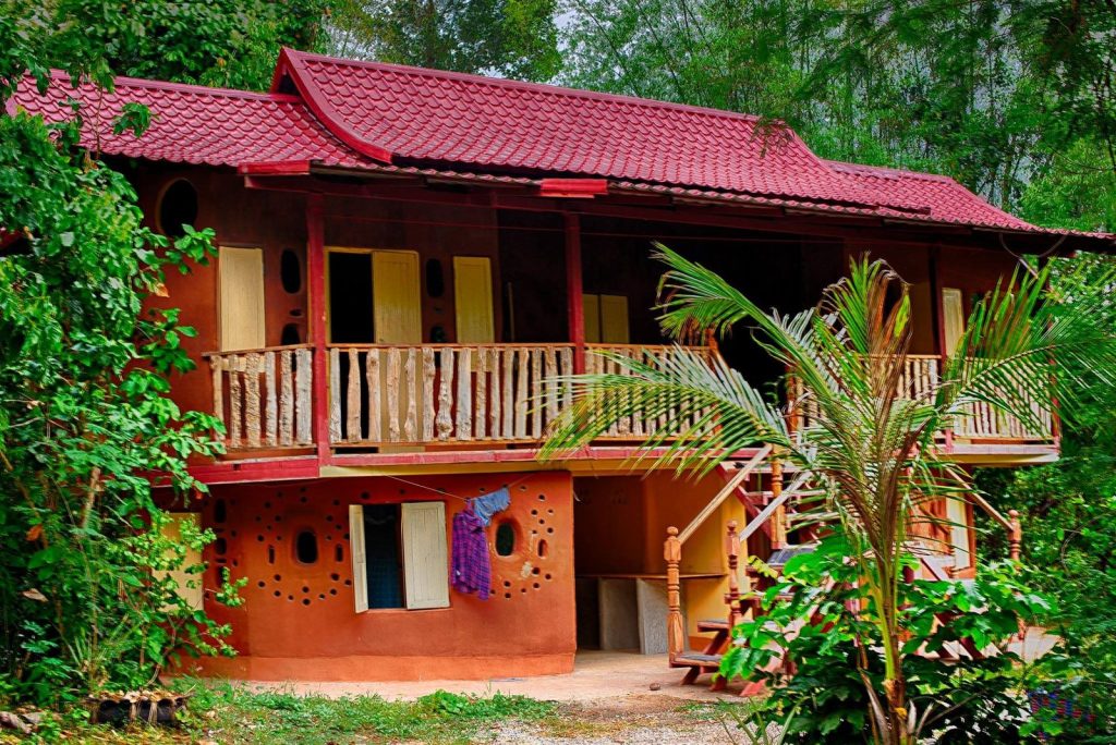 The Fairy House, a two-storey natural-built farm-stay cottage with a red roof and timber balcony, set among tropical greenery at Gaia Ashram.