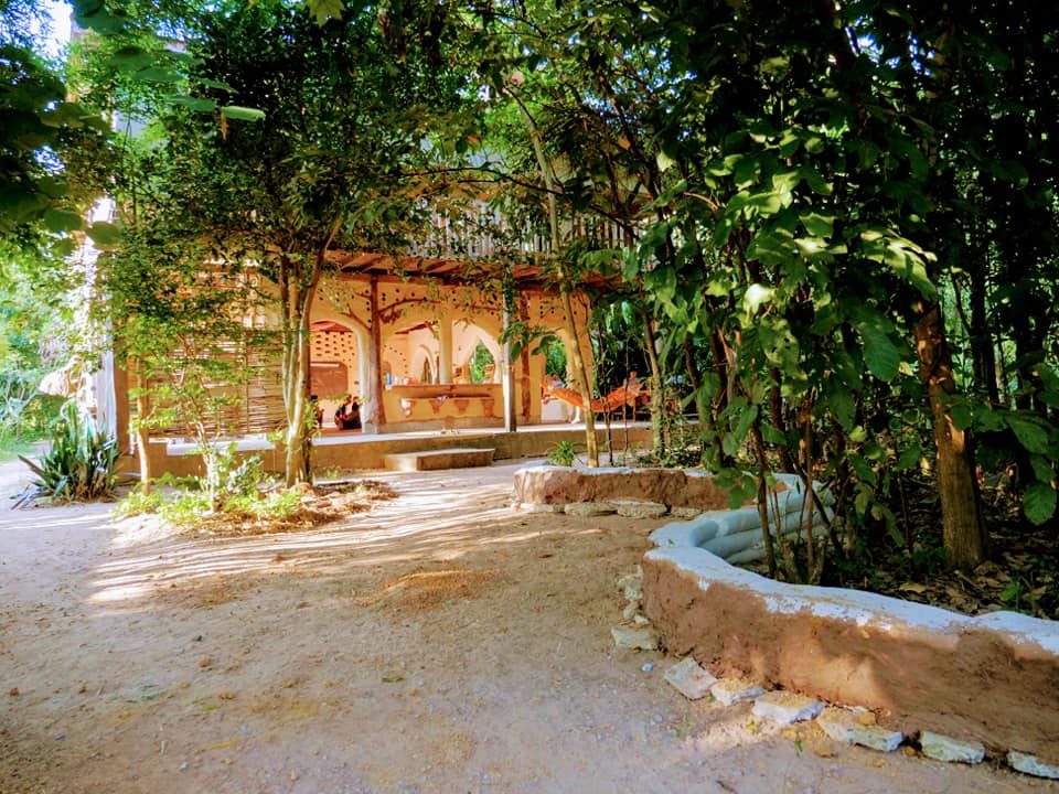 An open-sided timber and earthen sala set among trees, used as a learning space at Gaia Ashram.