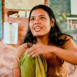 Om Sunisa Jamwiset Deiters, co-founder of Gaia Ashram, smiling and gesturing while teaching in front of a chalkboard.