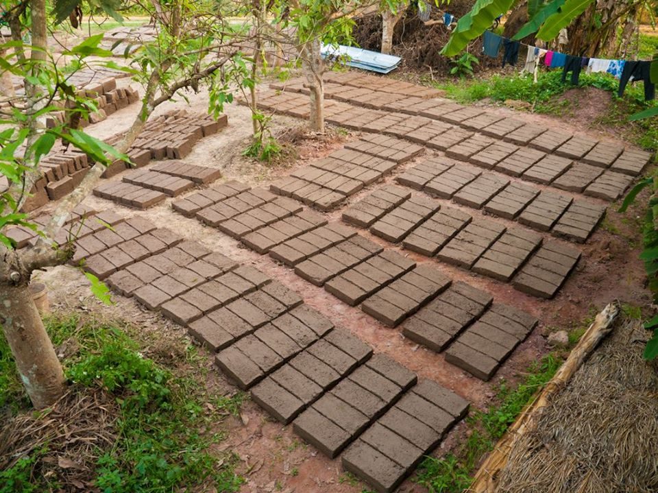 Rows of handmade mud adobe bricks laid out on red earth to dry in the sun among scattered fruit trees, with laundry on a line in the background, at The Panya Project, Mae Taeng, Chiang Mai.