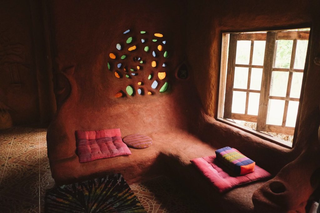 The interior of an earthen cob building with smooth sculpted reddish-brown walls, a built-in bench with Thai triangle cushions on a patterned tile floor, a wooden-framed window, and a swirl of coloured bottle-glass set into the wall to let light through, at The Panya Project, Mae Taeng, Chiang Mai.