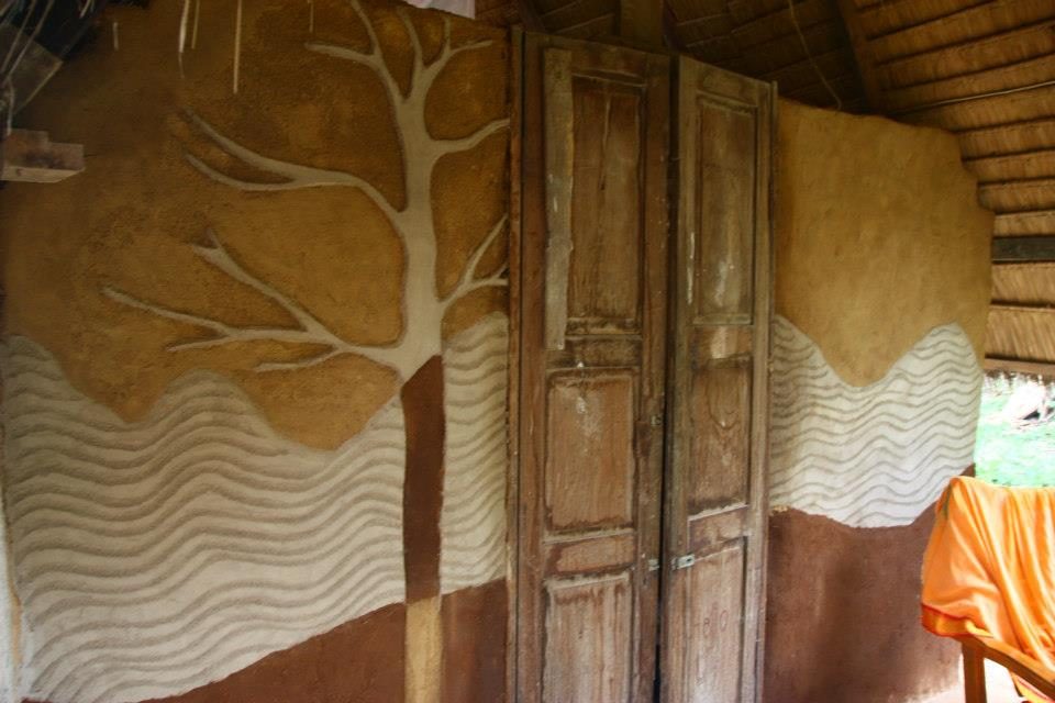 A natural-building wall decorated with a relief mural of a stylised tree in golden-brown earth over wavy textured plaster, beside a pair of weathered wooden doors, under a thatched roof, at The Panya Project, Mae Taeng, Chiang Mai.