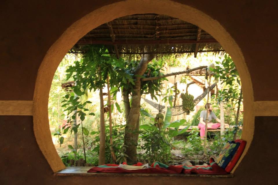 A round moon-shaped window cut into a smooth earthen wall frames a view out to a lush green food forest with banana and fruit trees and a person sitting on a raised platform under a thatched shelter, with colourful cushions on the windowsill, at The Panya Project, Mae Taeng, Chiang Mai.