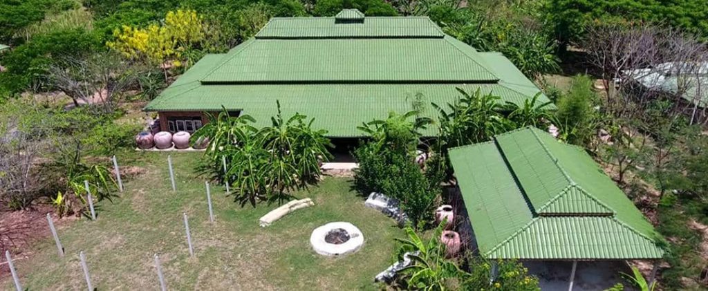 Aerial view of green-roofed farm buildings surrounded by fruit trees, banana plants and large terracotta water jars, at Rak Tamachat Permaculture, Si Khiu, Nakhon Ratchasima.