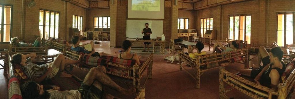 An instructor presenting to course participants reclining on bamboo daybeds in the brick-walled indoor classroom hall with a projector screen, at Rak Tamachat Permaculture, Si Khiu, Nakhon Ratchasima.