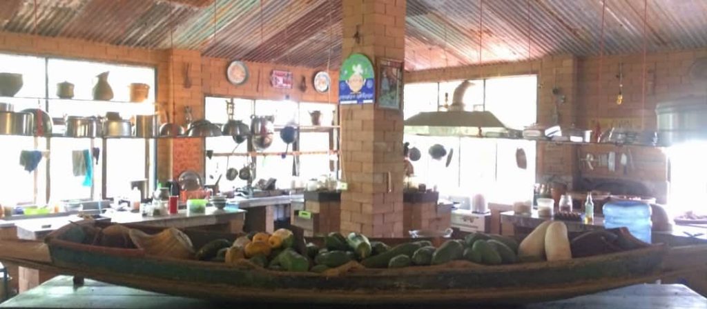 The open communal kitchen with hanging pots and pans and a wooden boat-shaped tray piled with squash and papaya in the foreground, at Rak Tamachat Permaculture, Si Khiu, Nakhon Ratchasima.