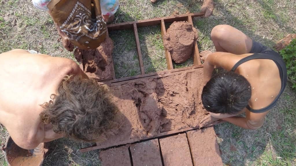 Overhead view of two people pressing wet earth into a wooden mould to make adobe mud bricks on the grass, with finished bricks drying nearby, at Rak Tamachat Permaculture, Si Khiu, Nakhon Ratchasima.