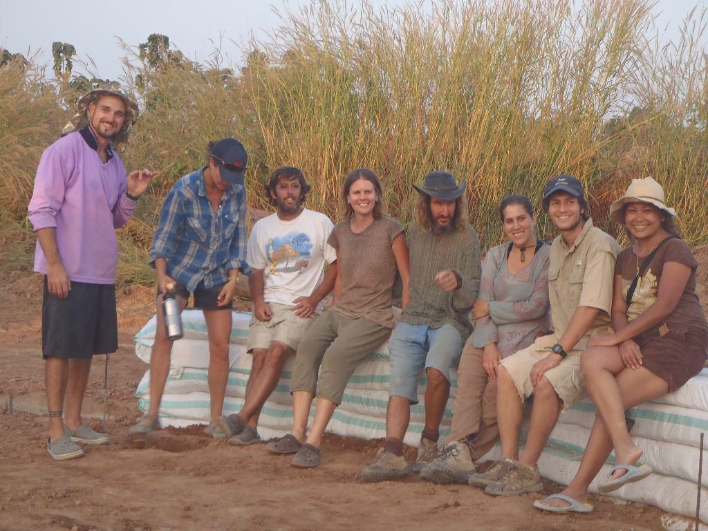 A group of natural-building course participants sitting together on a course of earthbag wall at dusk, with tall grasses behind, at Rak Tamachat Permaculture, Si Khiu, Nakhon Ratchasima.