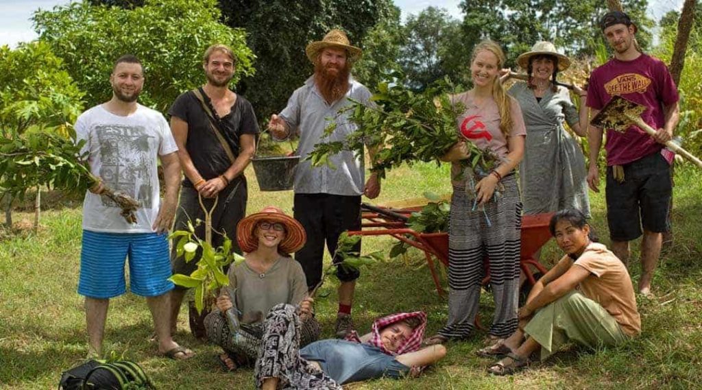 A group of course participants posing in the orchard holding bare-root tree seedlings and a wheelbarrow during a planting session, at Rak Tamachat Permaculture, Si Khiu, Nakhon Ratchasima.