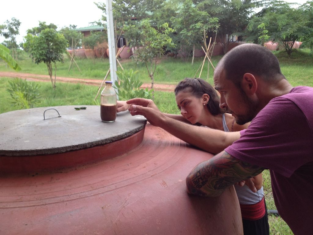 Two course participants examining a glass bottle of liquid resting on the lid of a large terracotta water jar, with farm buildings and a dirt track behind, at Rak Tamachat Permaculture, Si Khiu, Nakhon Ratchasima.