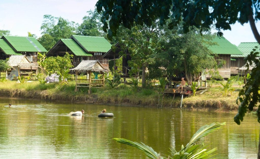 A row of green-roofed accommodation huts on the far bank of a pond, with people floating on inner tubes in the water, at Rak Tamachat Permaculture, Si Khiu, Nakhon Ratchasima.