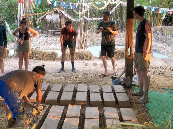 Guests making adobe mud bricks by hand under a bamboo shelter during a natural-building activity at Suan Na Ta Bua.