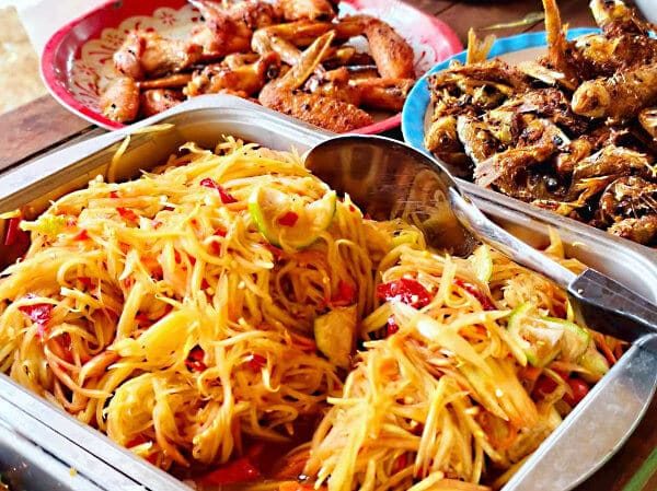 A tray of freshly made som tam (spicy green papaya salad) with plates of grilled fish and chicken, a farm-to-table Isaan meal at Suan Na Ta Bua.