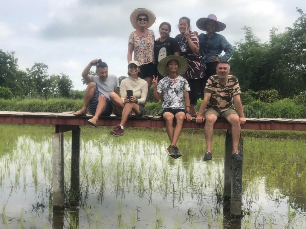 A group of guests and hosts sitting on a wooden boardwalk over a flooded rice paddy at Suwan Organic Farmstay, Phen, Udon Thani.