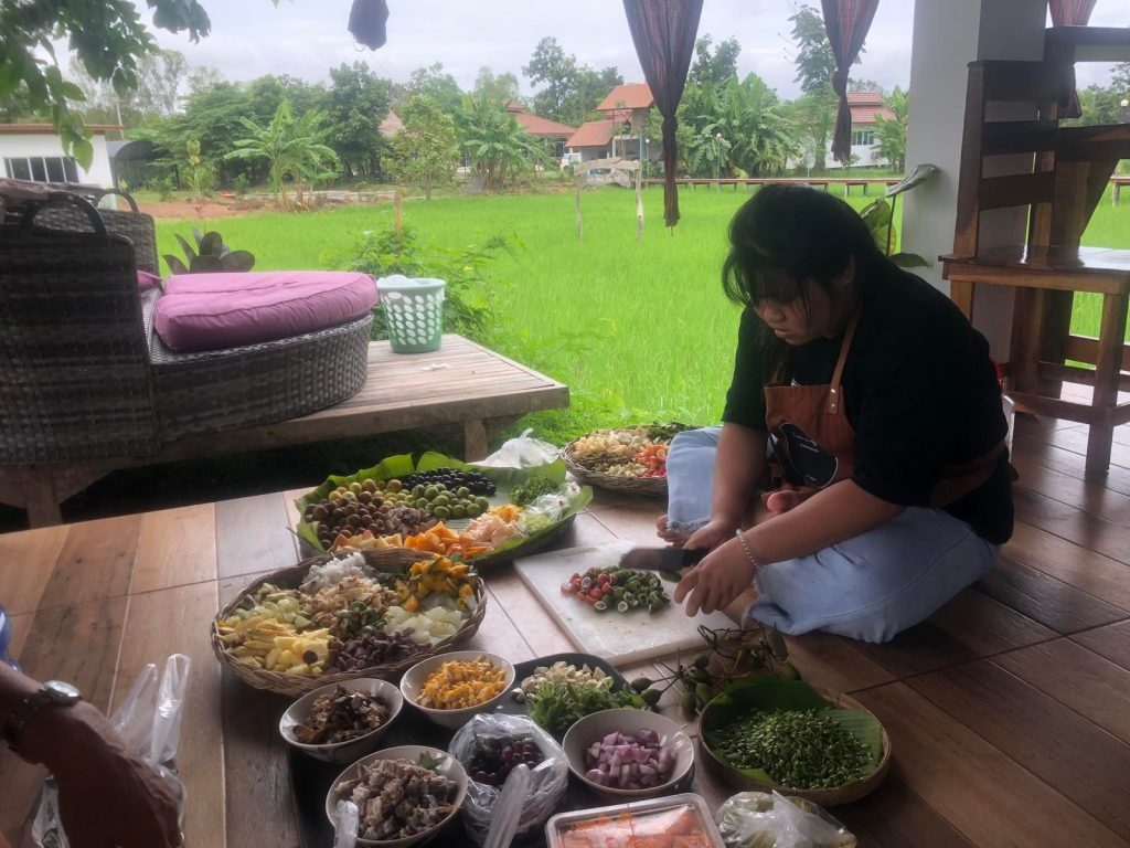 A cook preparing a spread of fresh vegetables and ingredients on the floor of an open terrace overlooking rice fields during a Thai cooking class at Suwan Organic Farmstay, Phen, Udon Thani.