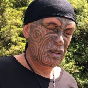 Te Kaha, Ngāi Tūhoe pounamu carver and storyteller, with full traditional facial tā moko, a black headscarf and a greenstone pendant, photographed outdoors by a river.