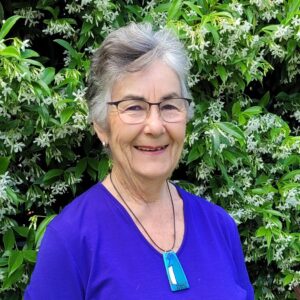 Trish Allen, permaculture practitioner from New Zealand, smiling in front of a flowering star-jasmine hedge.