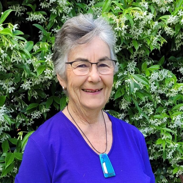 Trish Allen, permaculture practitioner from New Zealand, smiling in front of a flowering star-jasmine hedge.