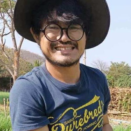 Nakorn Limpacuptathavon, Thai urban farmer, smiling in a wide-brimmed sun hat and round glasses in his vegetable field.