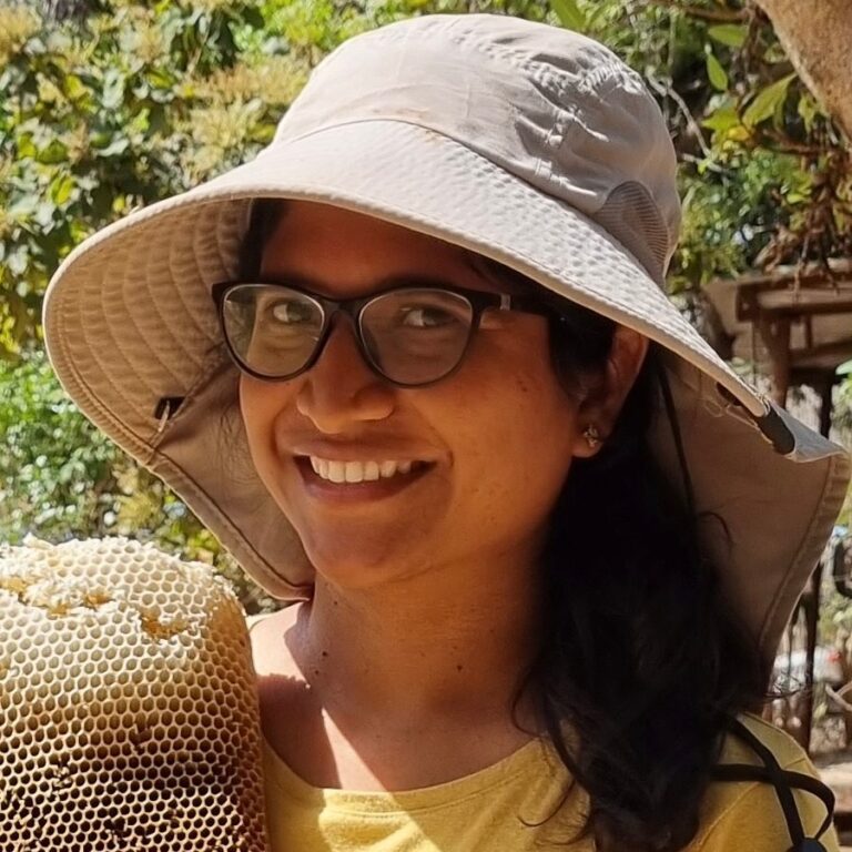 Sneha Koppula, permaculture educator, smiling in a wide-brimmed sun hat beside a frame of honeycomb at Aranya Permaculture Farm.