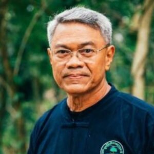 Wiwat Salyakamthorn, known as Ajarn Yak, a grey-haired Thai educator in glasses and a dark navy Thai-style shirt, standing in front of green forest.