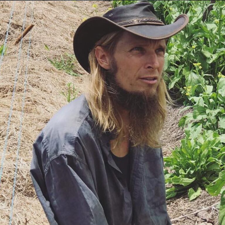 Bunya Halasz in a leather hat crouching beside a bed of flowering brassicas in a food forest at The Farm, Byron Bay.