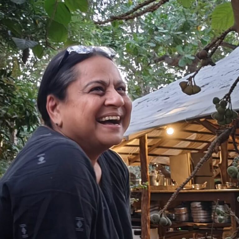 Clea Chandmal laughing beneath a fruiting tree at her farm in Goa, with an open-sided kitchen shelter behind her.