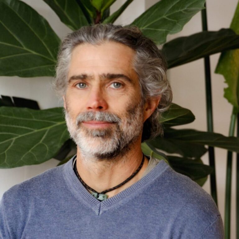 Finn Mackesy, with grey swept-back hair and a short beard, wearing a blue jumper and a pounamu pendant in front of large-leaved indoor plants.