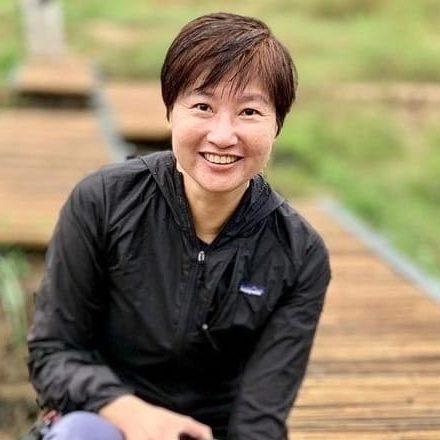 Idy Wong of Kadoorie Farm and Botanic Garden crouching on a bamboo walkway across a wetland, smiling.