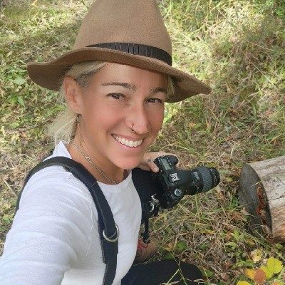 Lindsay Brandon in a wide-brimmed felt hat, smiling and holding a camera, kneeling on autumn grass.