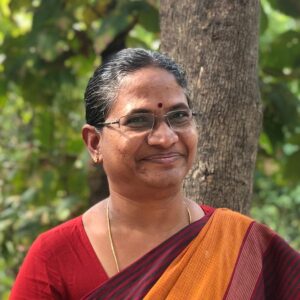 Padma Koppula, co-founder of Aranya Agricultural Alternatives, smiling in a red and orange sari in front of a tree.