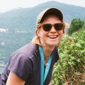 Paule de La Poype in a cap and sunglasses, laughing on a hillside path above the sea on Lantau, Hong Kong.