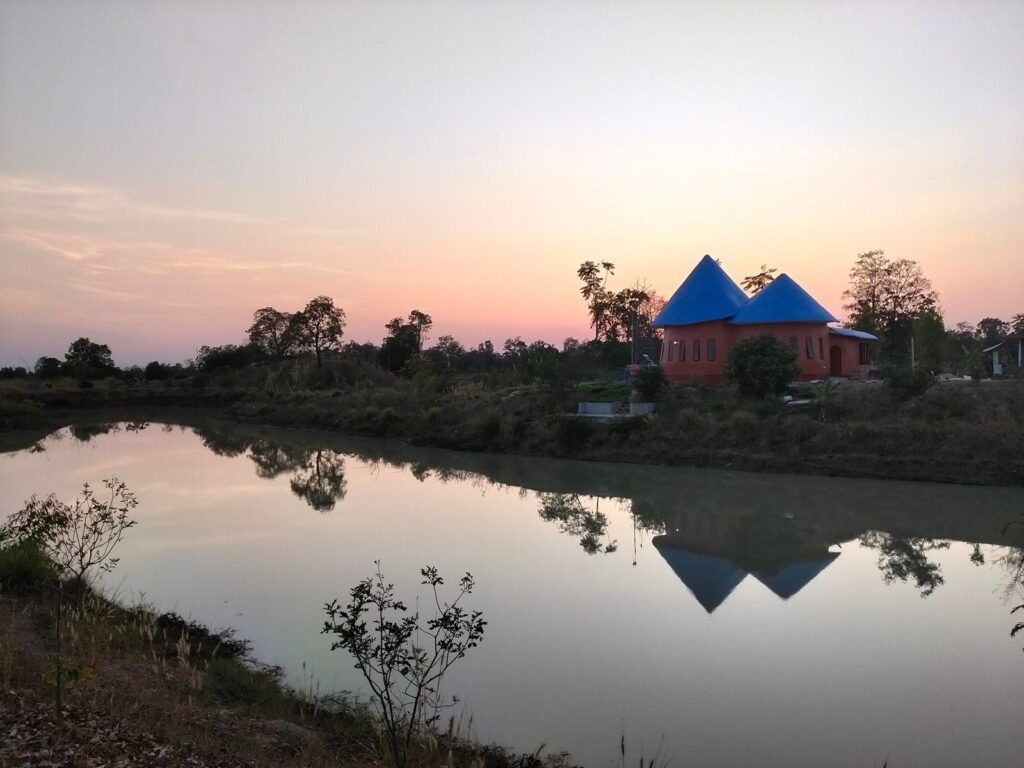 Sunset over the farm pond at Rainbow Mountain Permaculture Farm, Maha Sarakham, with a round orange house with two blue conical roofs reflected in the water.