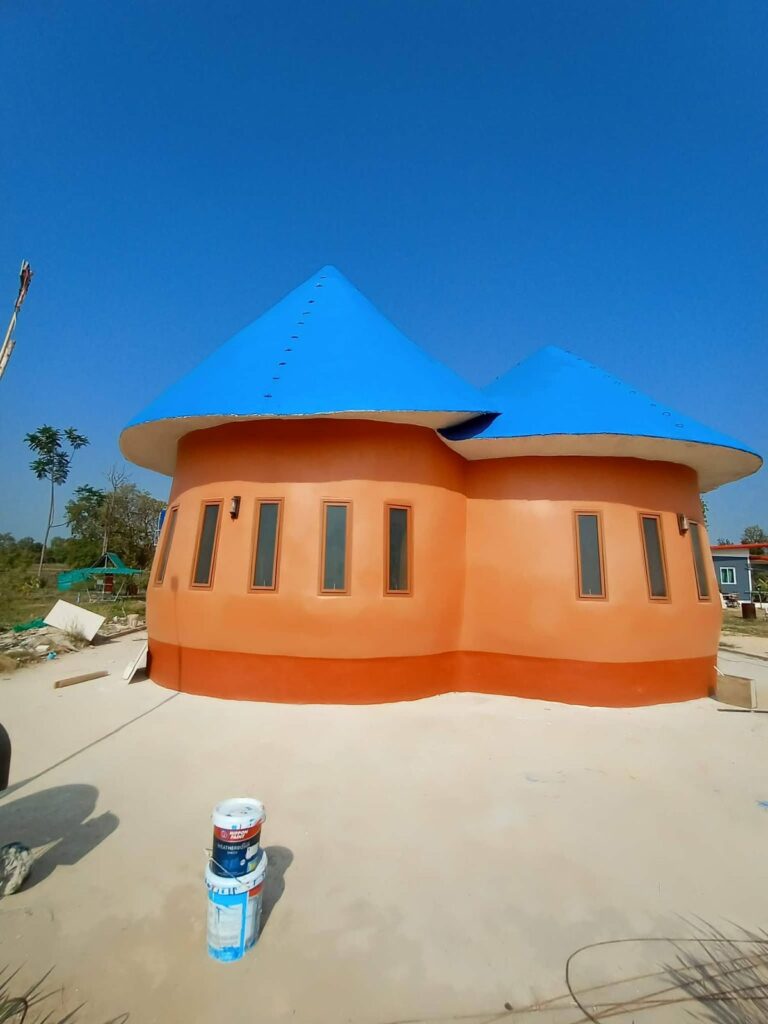 A round orange-plastered house with two blue conical roofs and narrow windows, newly built at Rainbow Mountain Permaculture Farm.