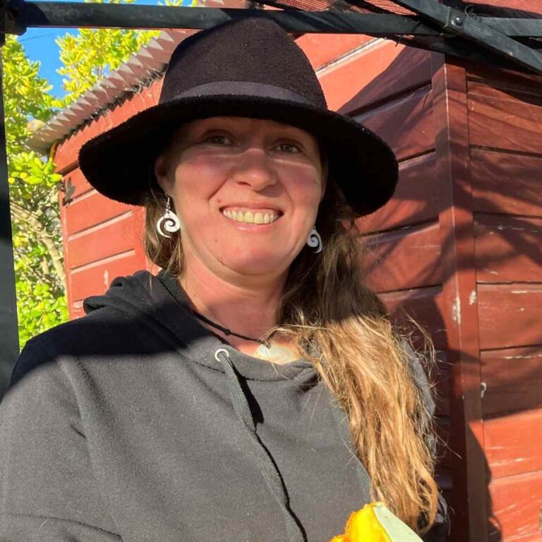 Roz Rolls in a black hat, smiling and holding a basket of harvested pumpkin and greens in front of a red shed.