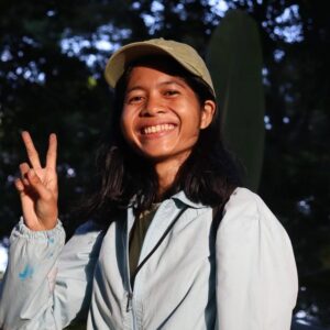 Sayu Komang smiling and giving a peace sign, in a pale cap and jacket under forest trees.