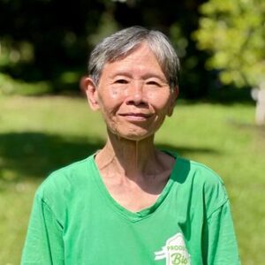 Darunee Tan, smiling, with short grey hair and a green T-shirt, standing on a sunlit lawn in front of trees.